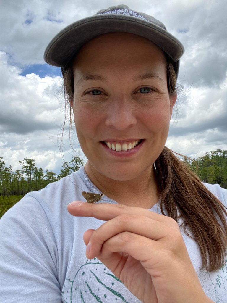 person smiling into the camera and holding a small butterfly on her index finger