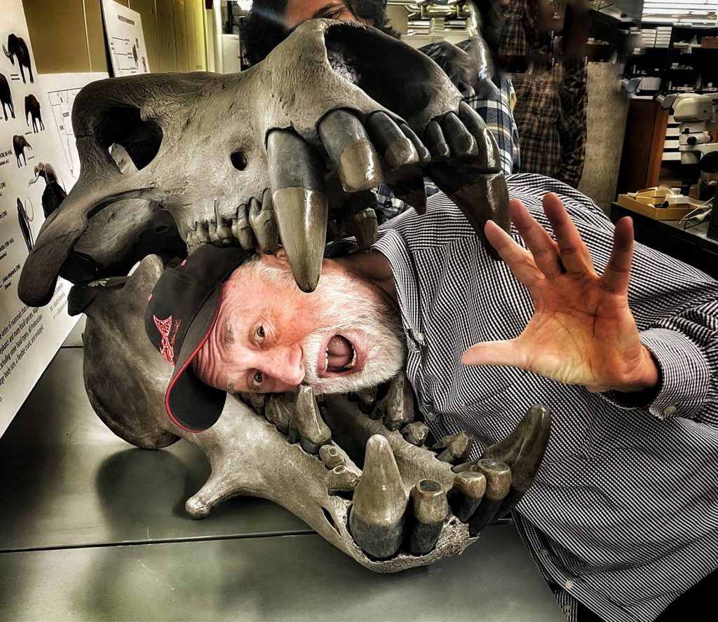 a man has put his head into the jaws of a large fossil skull with big teeth and is making a dramatic face