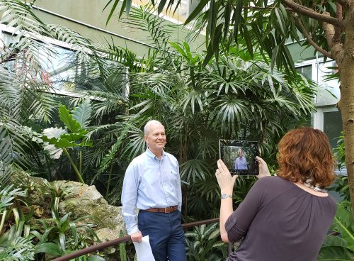 man standing in rainforest exhibit while woman films him with a tablet device