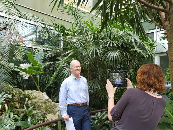 man standing in rainforest exhibit while woman films him with a tablet device