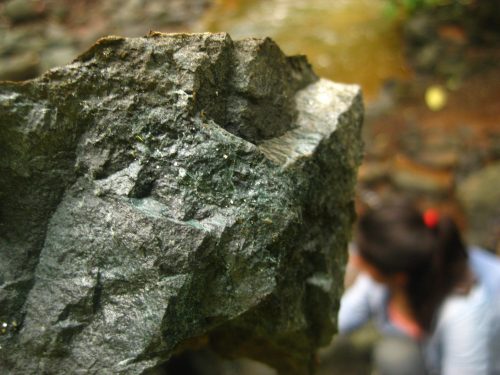 Tiny sparkling grains of a blue-green metamorphic mineral (most likely epidote) that forms when volcanic basalts are hydrothermally altered. Notice the thin orange layer (perhaps microbial) on the surface, which shows weathering or breakdown of the rock (‘meteorizado’ in spanish). Oftentimes it requires a rock hammer to break open the rock and see past this superficial rind.