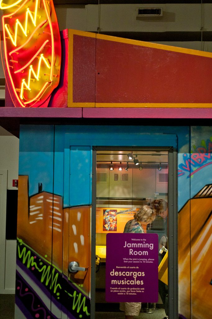 Glass door with the sign Jamming Room in English and Spanish. Through the door museum visitors can be seen exploring the exhibit