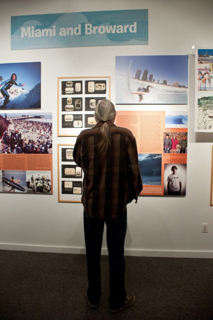 person with long hair stand in front of a wall filled with color and black and white photos of people surfing. All the images are under a sign that reads Miami and Broward