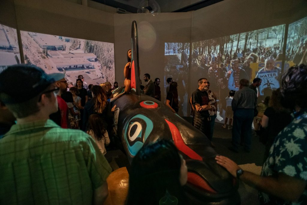 visitors walk around and touch the whale totem at the Whale people exhibit