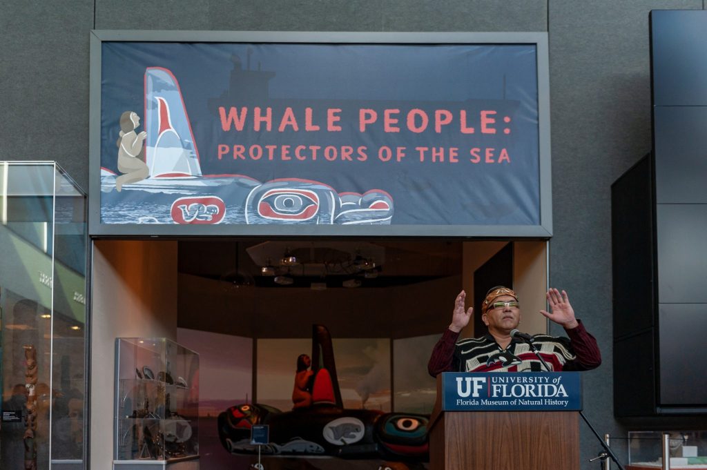 speaker stands at a podium in front of the entrance of the Whale People: Protectors of the Sea exhibit