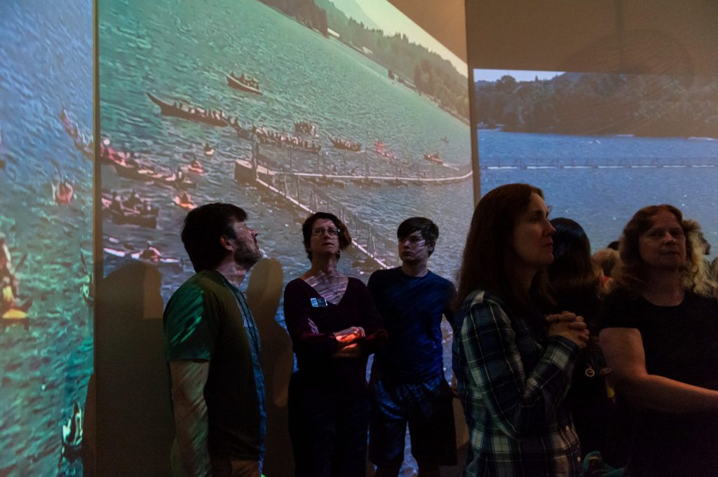 visitors look at the projected photos and videos on the walls of the Whale People exhibit