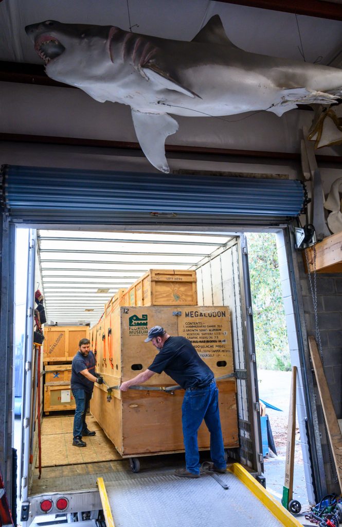 two men securing crates in a truck below a large fabricated shark