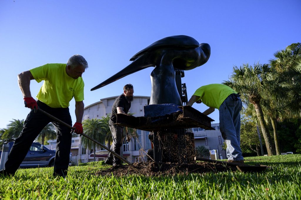 three men dig up the base of a large metal bird statue