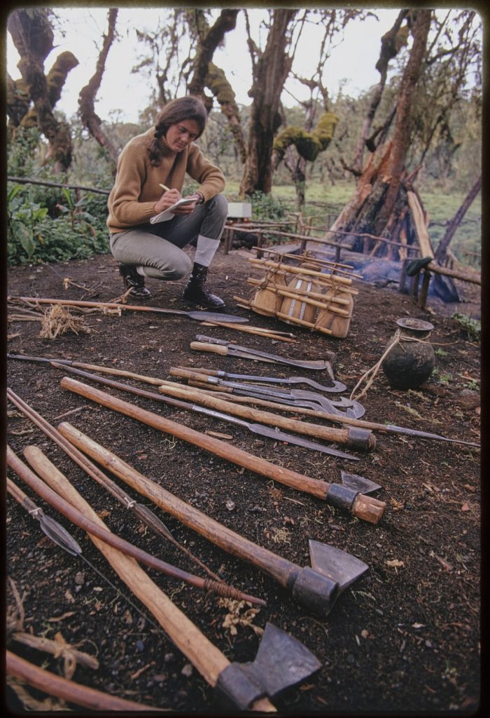 woman writes in notepad. on the ground around her are many axes and other weapons.