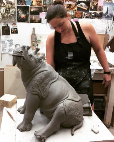 woman in apron covered in clay works on large clay sculpture