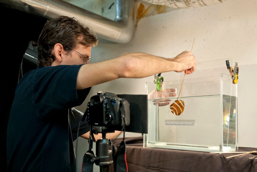 man placing a fish in clear tank filed with liquid. In front of the tank is a caner a on a tripod