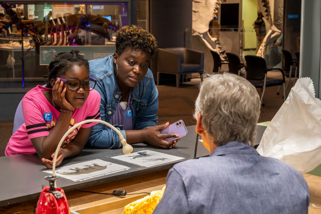 Two visitors, an adult and child watch as a researcher works in the Fantastic Fossil exhibit lab. The child is leaning forward to get a closer look.
