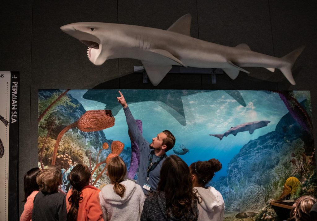 tour guild surrounded by children points to a model of a shark-like fish suspended from the ceiling