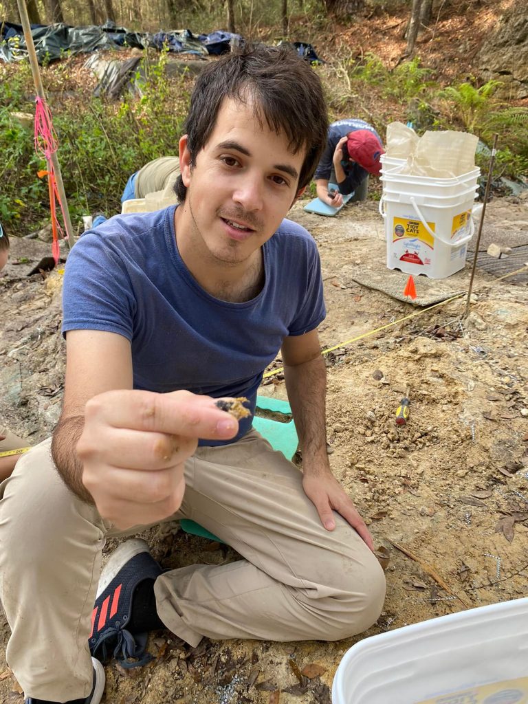 volunteer kneeling on the ground holding a fossil up to the camera