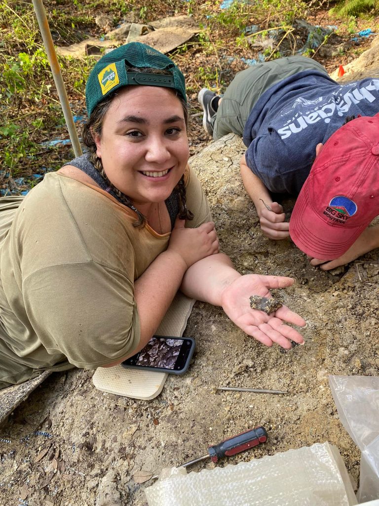 volunteers laying on ground holding a fossil and smiling at the camera