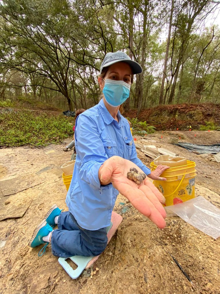 person wearing a mask kneeling on the ground holding out a hand with a fossil on it