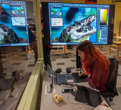 student works on computer, behind them a monitor shows a drawing of an alligator