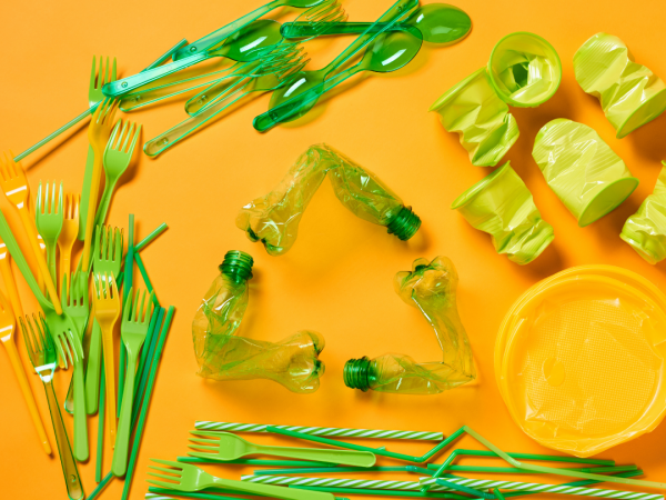 Plastic products including bottles, utensils, and cups formed to create a recycling symbol atop a bright orange background.