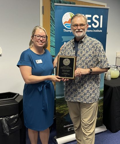 Ennes posing with MacFadden in front of the TESI vertical banner while they both hold the award plaque.