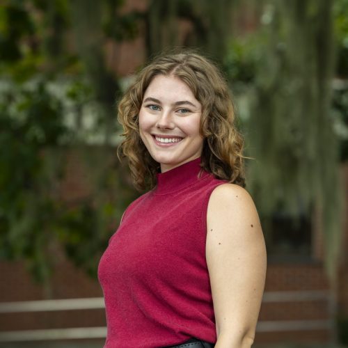 Hanley standing sideways and smiling at camera in front of greenery for headshot.