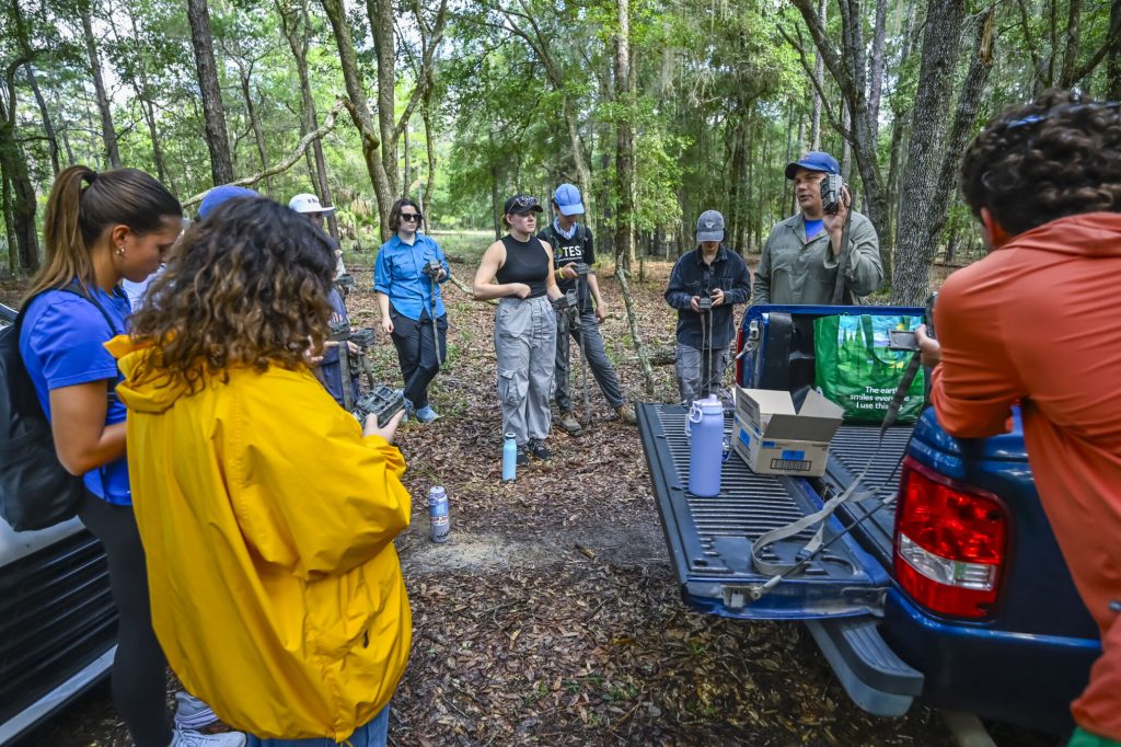Dr. Matt Hallett surrounded by the fellows by the cars at Ordway-Swisher as he explains how to turn on and set up a camera trap.