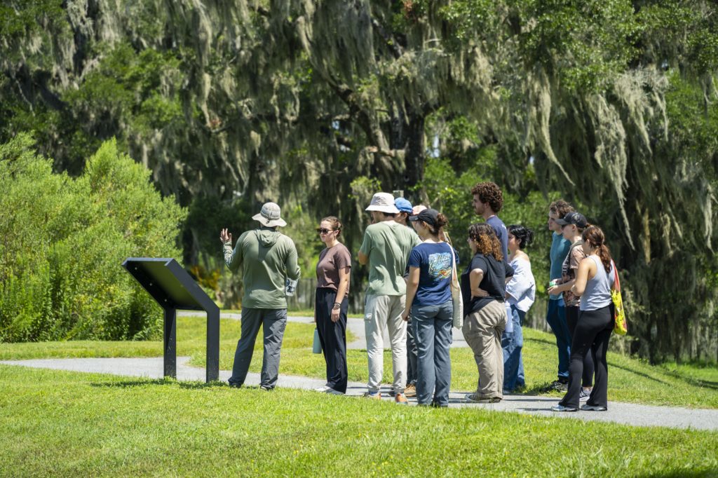 Fellows listening to Matthew Stanley at the Ocala Wetland Recharge park, as they stand in front of lark oak trees on the path.