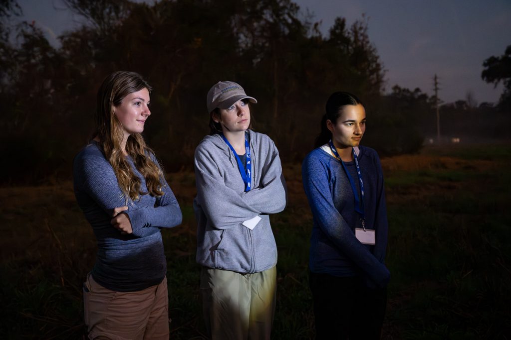 Left to right: Madison, Maggie, and Zoey listen to Charlie with light shining on their faces against the dark background.