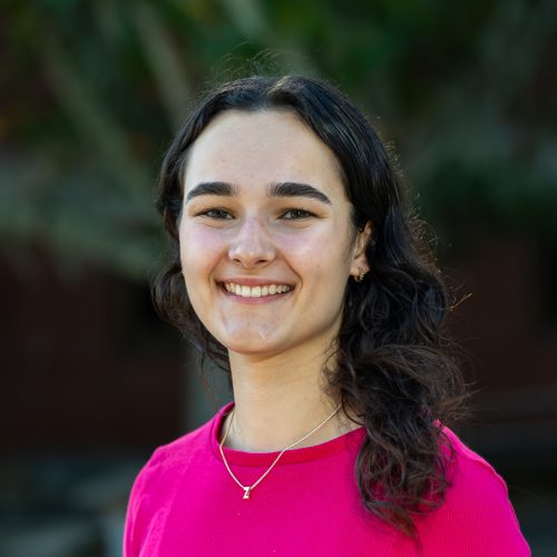 Zoey Papanikolaou wearing a hot pink shirt standing outside of Dickinson Hall and smiling for a headshot.