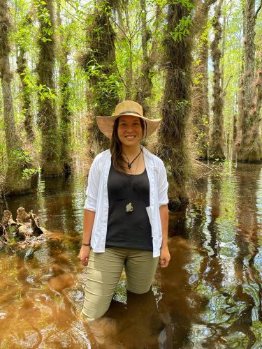 Woman standing in water up to her knees in the swamp.  She is wearing a wide brimmed hat and smiling.